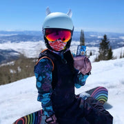 Child with small white horns mounted on his snowboarding helmet with snow in the background
