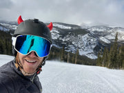 Man wearing small red horns on his ski helmet with snow in the background